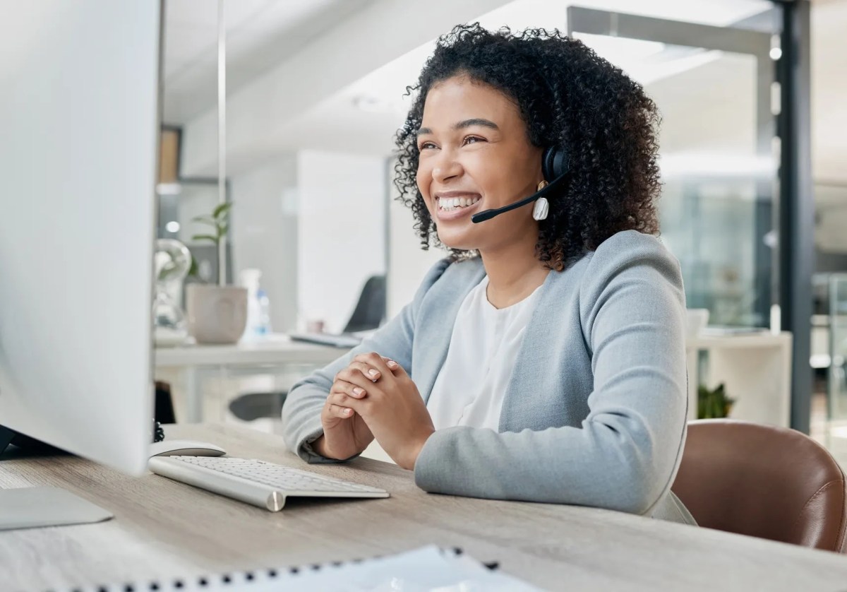A customer service representative smiles and listens to a call on a headset, while looking at a monitor.