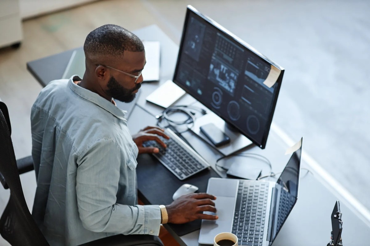 Looking down at an employee sitting at his desk using a desktop keyboard and large computer monitor and an open laptop.