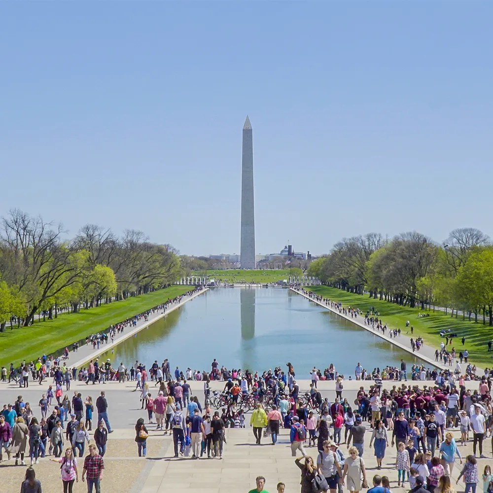 View of the Washington Monument and the Lincoln Memorial Reflecting Pool on a clear sunny day with crowds of D.C. residents and tourists.